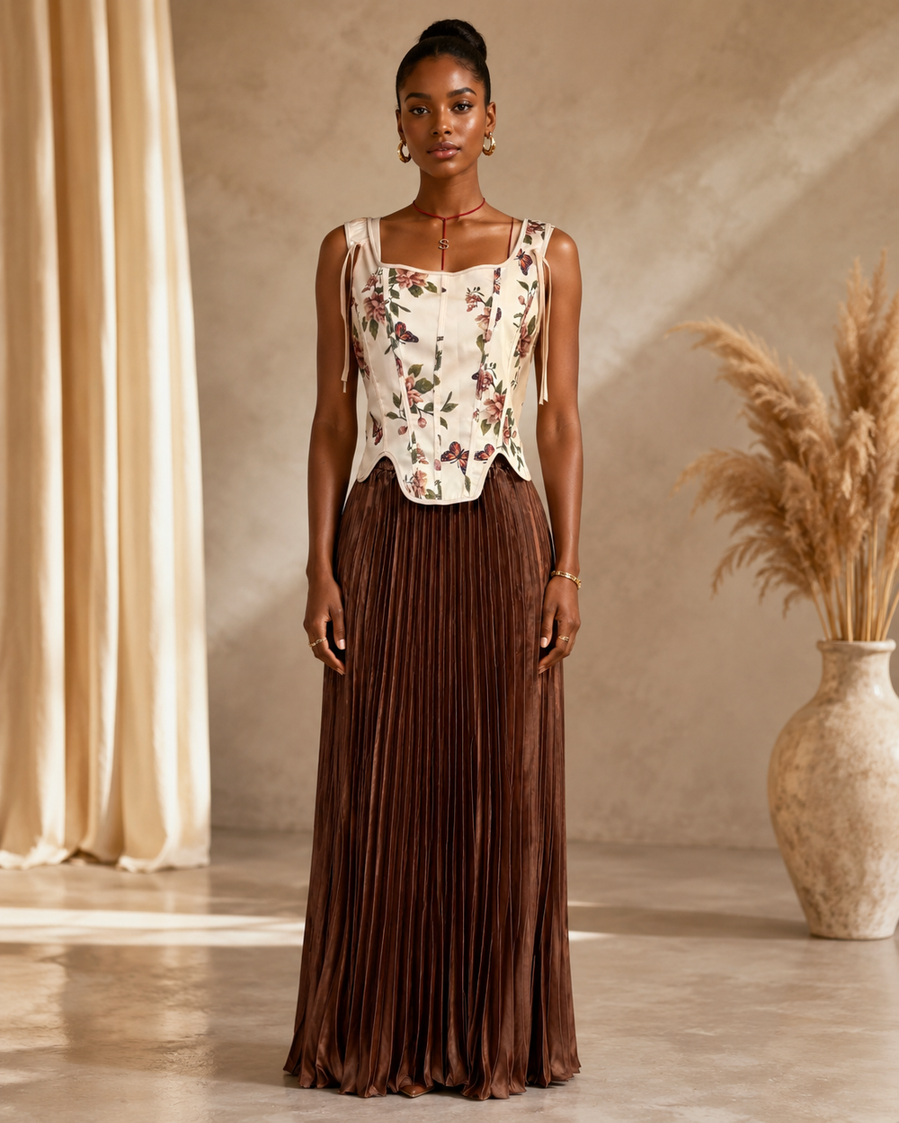 A woman stands in a warm, neutral-toned room wearing a sleeveless cream top with a floral print and a long brown pleated skirt.