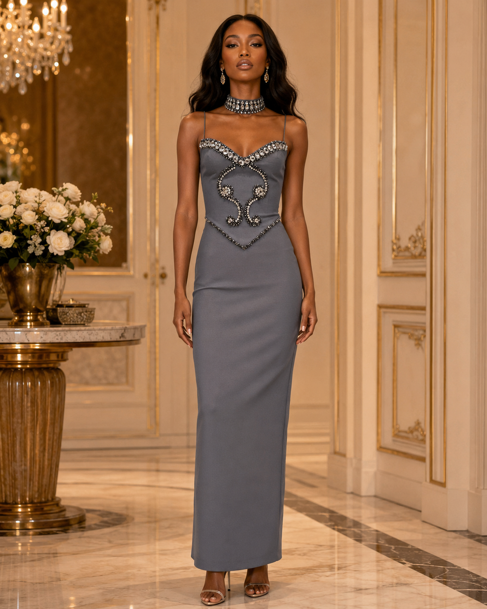 A woman stands in an elegant indoor hallway wearing a floor-length strapless gown in a muted blue-gray color with ornate silver embellishment across the bust and waist. She is wearing a matching silver choker and clear high-heeled sandals.