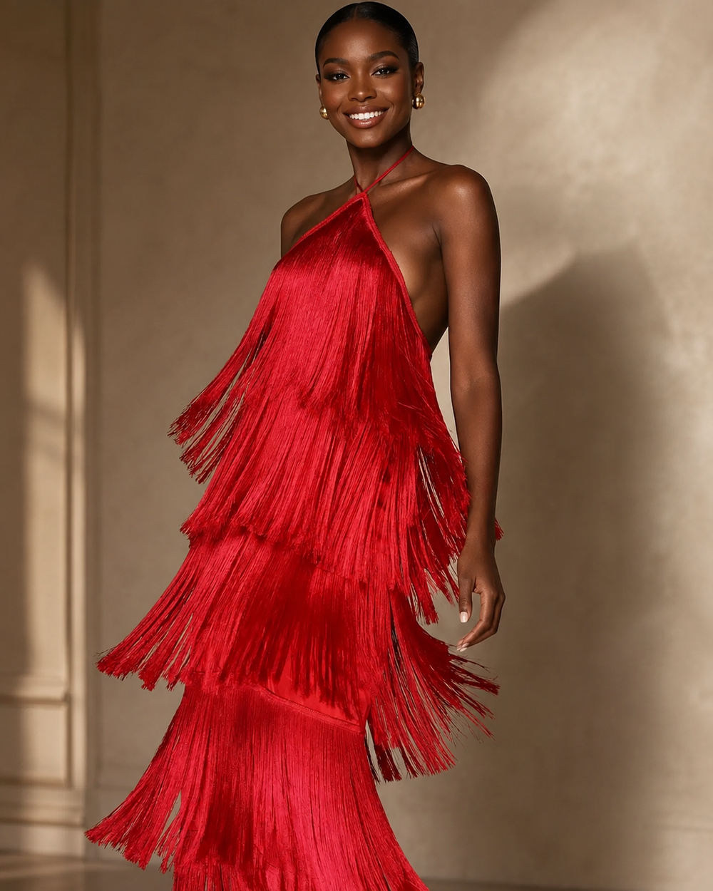 A woman modeling a vibrant red halter-neck dress with dramatic, layered fringe cascading down the front, set against a warm beige wall with soft shadows.