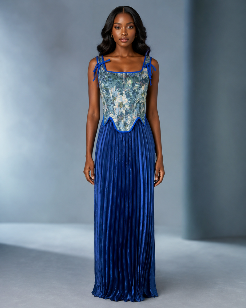 A woman modeling a sleeveless floor-length gown with a blue floral embroidered bodice and a pleated royal blue skirt, photographed against a soft gray studio backdrop.